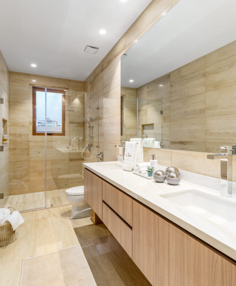 Baño moderno con ducha de vidrio, doble lavamanos, espejo amplio y acabados tipo piedra en The Woods Santa María.