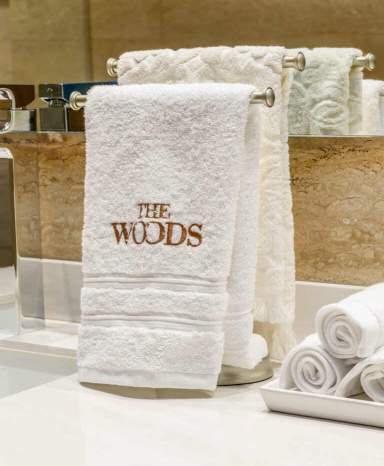 Toalla blanca bordada “The Woods” en baño, junto a toallas enrolladas y lavamanos.